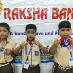 Three young schoolboys in beige uniforms holding colorful crafts at a school event standing in front of a banner